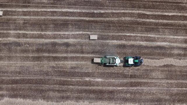 An aerial view shows a tractor pulling a baler, making straw bales in a vast agricultural field during the harvest season.