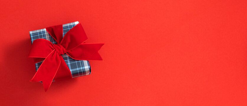 Christmas Gift Box with Red Velvet Ribbon Bow on Red Background Panoramic Banner. Plaid Present Left Side Flat Lay with Copy Space. Minimalist New Year, Holiday Greeting Card Mockup Concept.