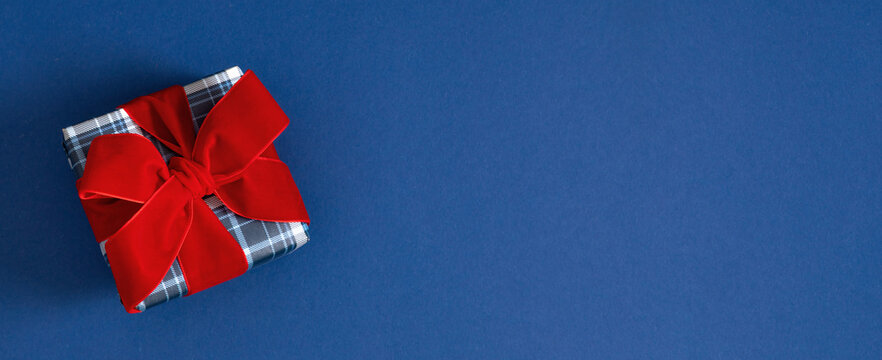 Christmas Gift Box with Red Velvet Ribbon Bow on Dark Blue Background Panoramic Banner. Plaid Present Left Side Flat Lay with Copy Space. Minimalist New Year, Holiday Greeting Card Mockup Concept