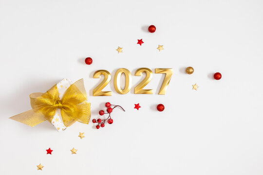 Happy New Year 2027 Festive Flat Lay Background with Golden Numbers. Christmas Gift Box with Ribbon, Red Berries and Confetti Stars on White Backdrop. Holiday Greeting Card Concept with Copy Space.