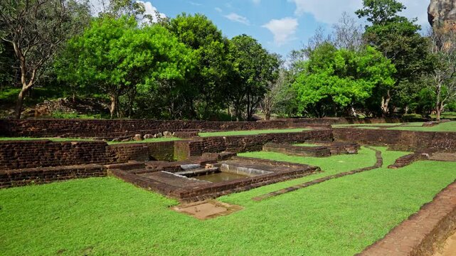 Sunlit brick terraces and walls of the ancient garden complex at the base of Sigiriya rock, with lush green lawns and trees under a blue sky.