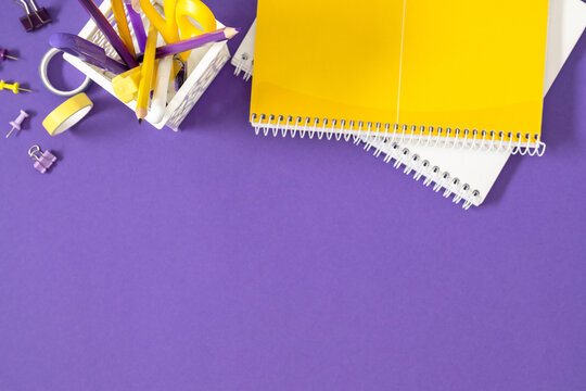 Yellow and Purple School Office Stationery Top Border Flat Lay with Copy Space. Spiral Notebooks, Pencils, Scissors in Holder on Violet Background Top View. Creative Minimalist Workspace Concept.