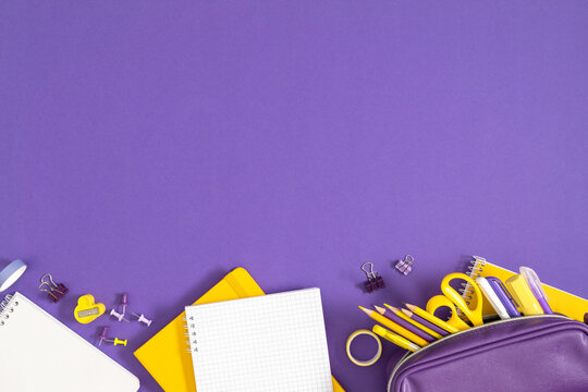 Purple Pencil Case and Yellow Stationery Bottom Border Flat Lay with Copy Space. Open Grid Notebook, Scissors, Pencils and Clips on Violet Background Top View. Creative Back to School Concept.