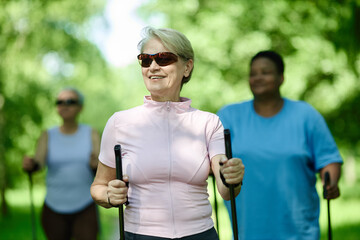 Naklejka premium Senior Caucasian woman smiling during Nordic walking with diverse group in park, promoting active aging and senior fitness. Image suitable for healthcare and retirement lifestyle marketing