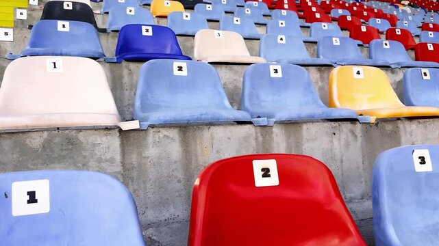 Close-up of numbered plastic stadium seats in red, blue, white and yellow, with concrete rows and bright yellow stairs in an empty sports venue.