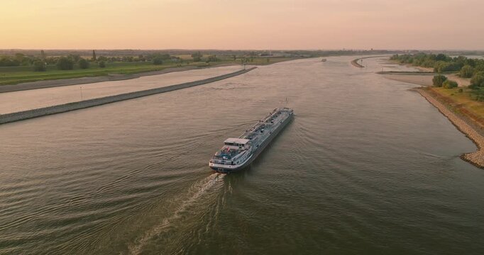 Drone orbits behind a cargo ship sailing through a Dutch canal at sunset, revealing the vessel's wake, tranquil water, colorful skies, and the iconic Dutch polder landscape.