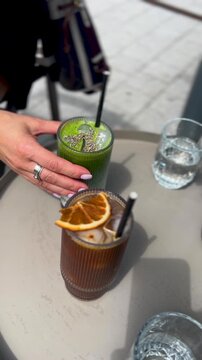 A close up vertical shot of a womans hand reaching for healthy iced drinks including matcha and cold brew at an outdoor cafe. Authentic lifestyle scene for social media and wellness content.