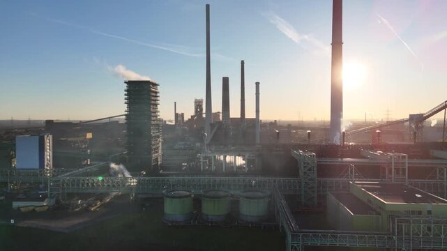 Drone passes industrial chimneys with parallax as morning sun hides behind a front stack