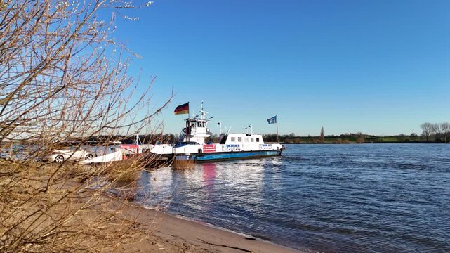 Drone rises above foreground shrubs as vehicles drive onto the ferry, revealing river traffic from an elevated boarding perspective.