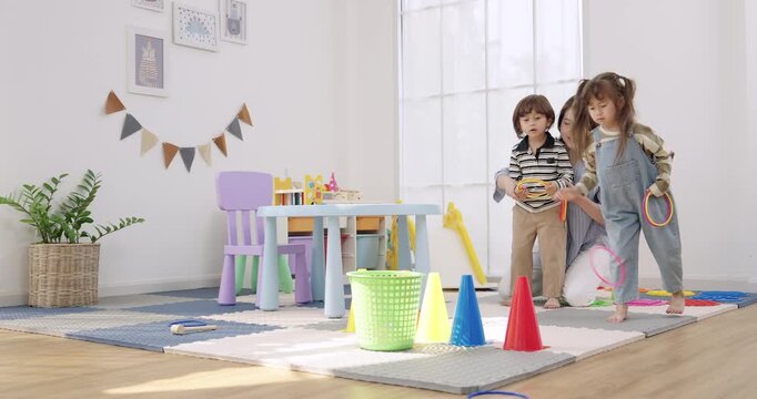 Asian female preschool teacher watches toddler boy, toddler girl display ring set beside basket, cone course on floor, kindergarten game promotes sharing, confidence, social interaction, childcare