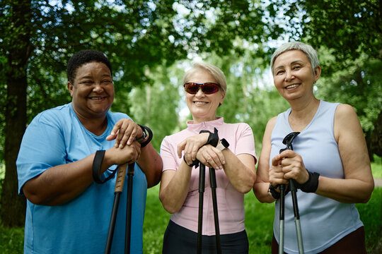 Portrait of diverse senior women smiling during nordic walking in park, promoting active lifestyle. Image suitable for healthcare campaigns and senior wellness marketing, medium shot