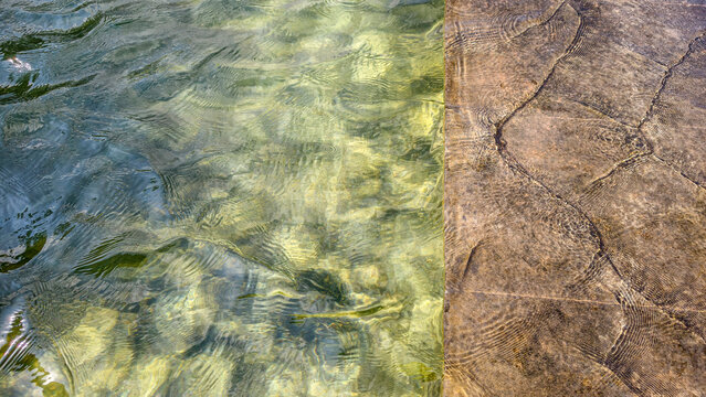 Rippling water reflecting sunlight on clear pool surface, flowing over submerged beige paving stones, creating an abstract texture