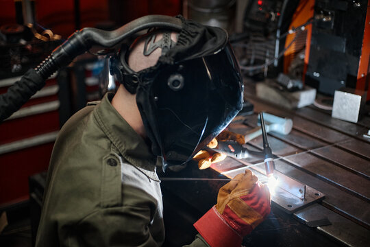 Caucasian male welder wearing protective mask and gloves welding metal in workshop, representing industrial manufacturing and vocational training, high angle shot