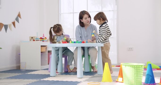 Asian female preschool teacher assists toddler girl, toddler boy sorting rings and cones on play table, daycare classroom routine, color learning, teamwork, creative education support, fine motor