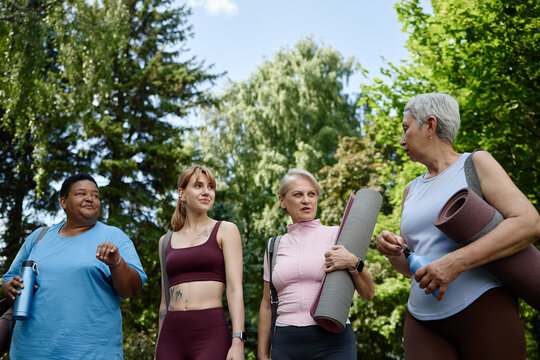 Diverse group of multiethnic women walking in park with yoga mats, talking after outdoor workout. Low angle shot representing active lifestyle, wellness, and community fitness marketing