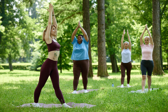 Diverse multiethnic women practicing yoga in park, stretching arms up on mats. Image represents body positivity and wellness, suitable for fitness and mental health marketing campaigns