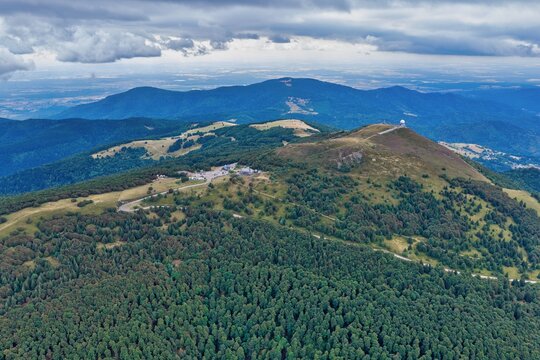 Aerial view of the Grand-Ballon mountain featuring its distinctive radar dome, winding mountain roads, and dense forests under a cloudy sky in Soultz-Haut-Rhin, Grand Est, France.