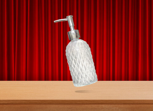 A clear glass perfume bottle with a diamond pattern sits on a wooden table, with a red curtain as a backdrop.