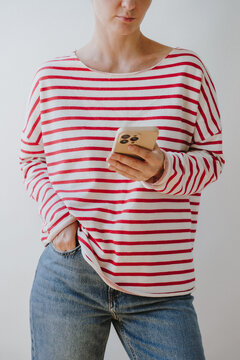 Woman Wearing Red And White Striped Long Sleeve Shirt Holding Smartphone Standing Against Plain Wall