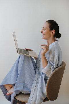 Side View Of Laughing Woman Sitting On Chair Having Video Call On Laptop In Light Blue Shirt