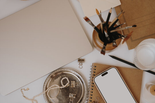 Top View Cozy Artist Workspace Setup Featuring Blank Mobile Phone Screen Mockup, Kraft Paper Journals, And Paintbrushes