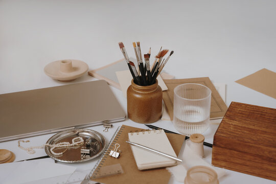 Aesthetic Earth Tone Desk Setup With Closed Laptop, Paintbrushes In Clay Pot, And Kraft Notebooks