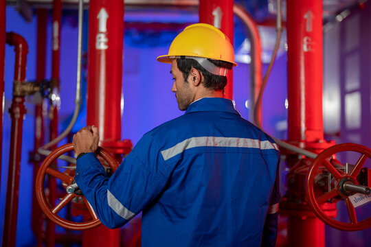 A male engineer in a blue coverall and hard hat operating the valve wheel by the red piping in a fire pump room with red piping and valves. An authentic industrial mood.