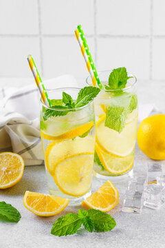 Close up of two clear glasses with refreshing homemade lemonade with fresh lemon slices, mint leaves, ice cubes and paper straws on a light kitchen counter against white tile background