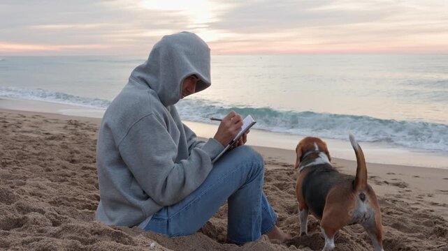 Peaceful moment of a creative writer in a hoodie sitting on the sand and taking notes in a journal at sunrise, accompanied by a loyal beagle dog enjoying the calm ocean waves and scenery