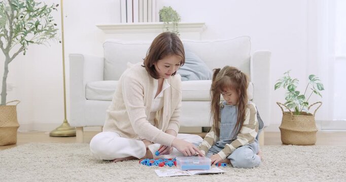 Asian single mother sliding blocks across carpet, watching preschool daughter assemble tower, cheering success, practicing sharing skill through cooperative play, indoor playtime learning livingroom.