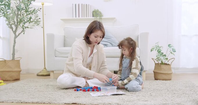 Asian single mother kneeling on carpet, sorting toy blocks into box, pointing toward worksheet, guiding preschool daughter following steps, practicing math skill, indoor livingroom learning.