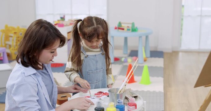 Slow motion clip: adult asian mother teaches preschool daughter dipping brush, mixing pigment on palette, then painting easel canvas, weekend homeschool routine, calm bonding, child development.