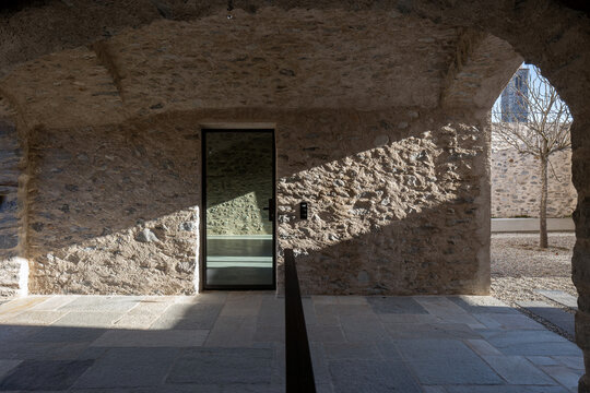 The covered entrance to an old house that has been completely renovated, with stone walls. A beam of light streams through a large arch onto the wall&rsquo;s surface. In the centre is a glass door. 