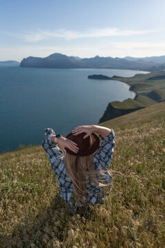 Woman traveler hiking, sitting on green grassy hill overlooking scenic coastal mountains and blue sea during summer adventure trip