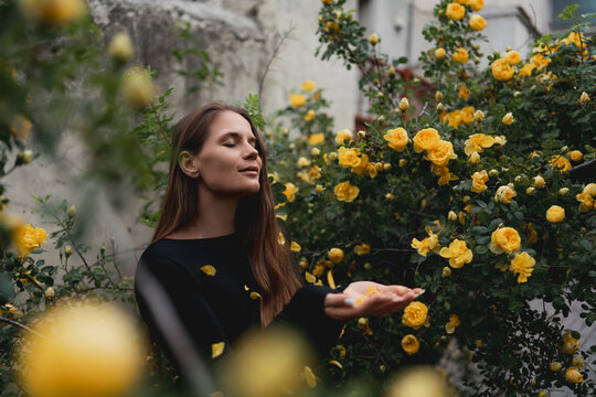 Young woman enjoys falling petals of yellow roses while standing in a lush summer garden