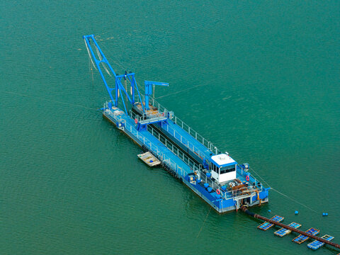 Aerial view of a blue sand mining dredger operating on calm water with a floating pipeline and a small support boat in Poland.