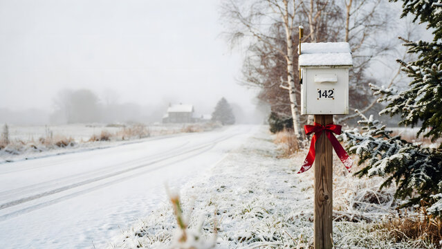 A white mailbox with the number 142 and a red ribbon on a wooden post stands beside a snowy road in a cold, overcast winter rural landscape