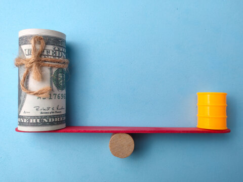 A roll of US dollars balancing against a miniature yellow oil barrel on a red wooden seesaw over a blue background