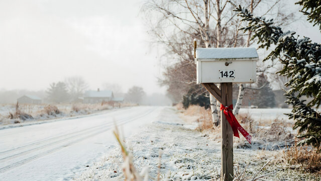 A white metal mailbox with the number 142 stands on a wooden post decorated with a red ribbon along a snowy road in a quiet winter landscape