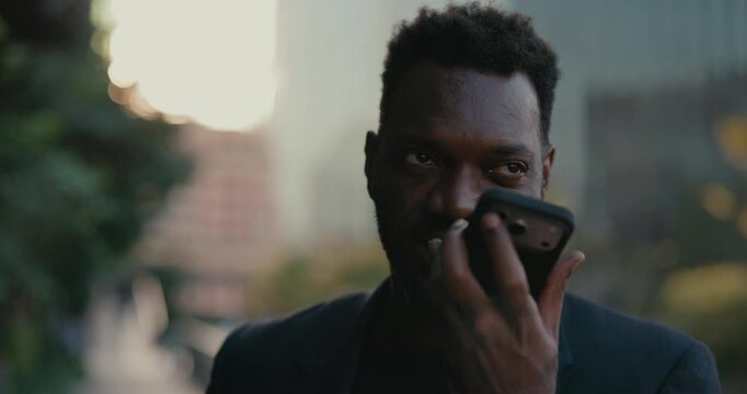 African American businessman records a voice message on his smartphone outside a modern office, speaking clearly while sending mobile audio communication to a colleague.