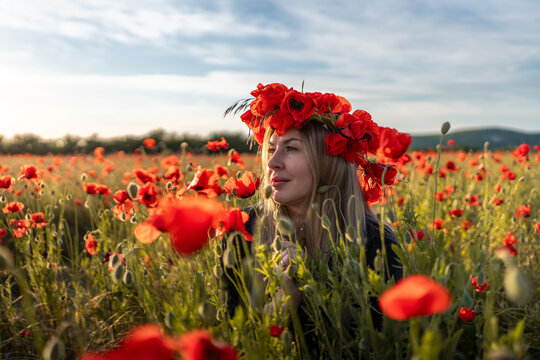 Blonde woman wearing Papaver rhoeas flower crown stands in sunny summer field of red poppies