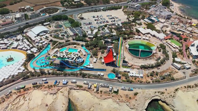 Baħar iċ-Ċagħaq, Malta aqua park with waterslides, pools, wave pool and lazy river beside the Mediterranean Sea, families and children enjoying leisure activities, drone view.