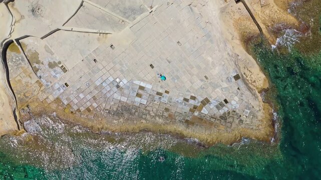 Baħar iċ-Ċagħaq, Malta salt pans and rocky Mediterranean shoreline with geometric patterns, blue water, and people swimming and sunbathing, captured by drone in summer.