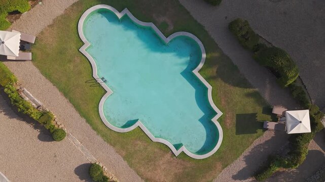 Drone ascends over ornate swimming pool with sun loungers and umbrellas in garden setting