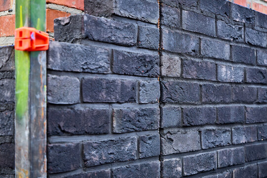 Dark clinker brick wall under construction showing a vertical expansion joint, mason's corner guide profile, and orange line block clamp with alignment string.