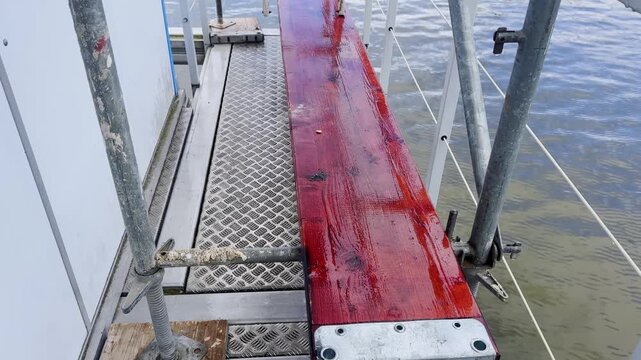 Floating platform walkway with a wet red wooden plank, metal grating floor, scaffold poles, railings and lake water beside a temporary event structure.