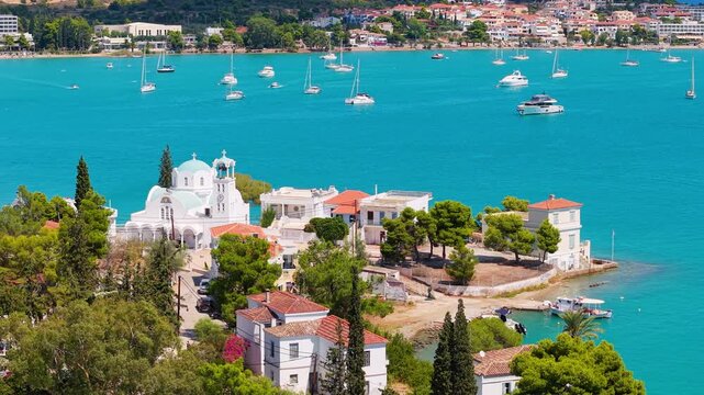 Drone pans over an iconic Greek Orthodox church with blue dome on the Porto Cheli waterfront. Turquoise bay, sailboats and the village shimmer under summer sun.