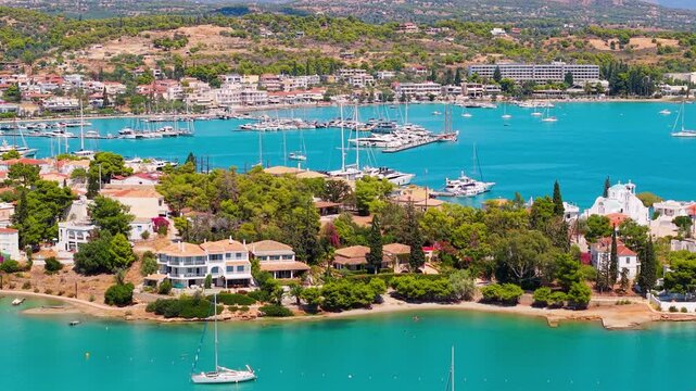 Drone pans over Porto Cheli revealing its charming village, packed marina and turquoise bay. Luxury yachts, white villas and a church shine under the summer sun.