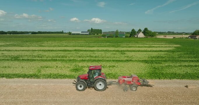 Red tractor producing straw bales on harvested farmland in a peaceful sunny countryside landscape with green fields and a clear blue summer sky, showcasing modern agricultural work.