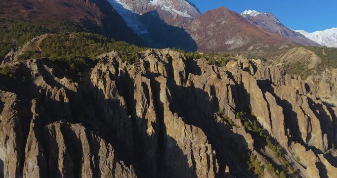 Annapurna Circuit, Manang, Nepal, drone shot over dramatic eroded cliff formations and pine forest revealing snow capped Himalayan peaks on a bright sunny day.